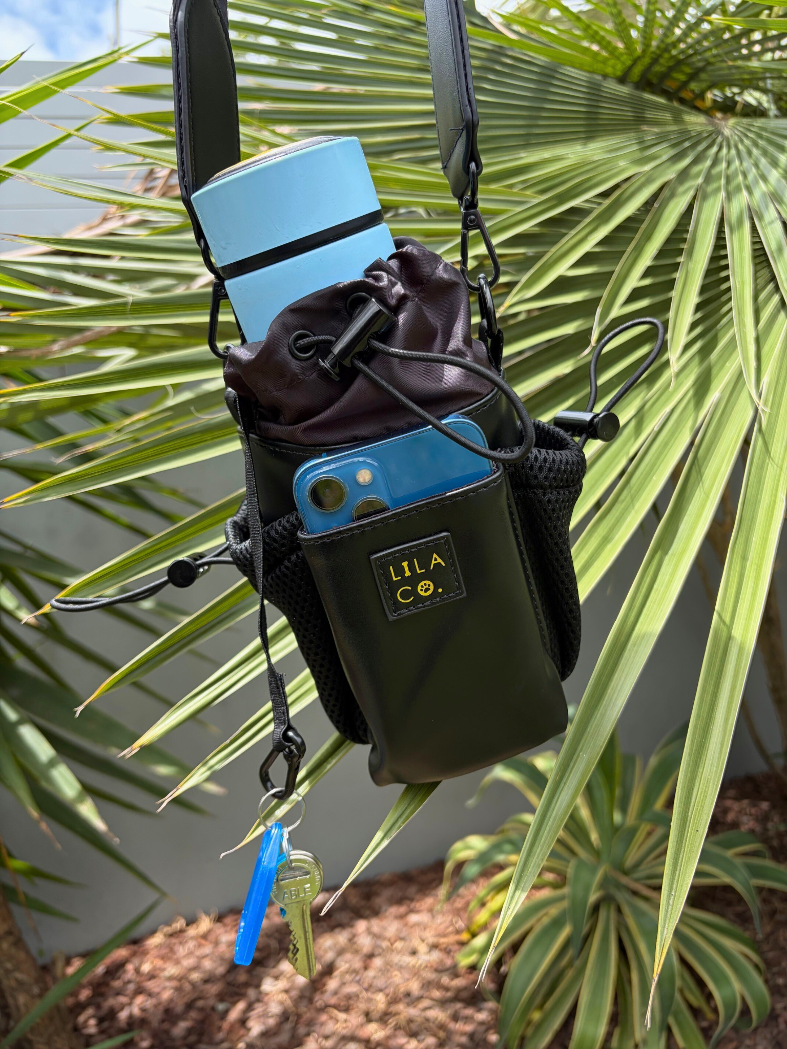 Black pouch with blue accents and 'Lila & Co.' branding hanging on a palm leaf.