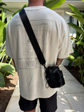Person wearing a white outfit with a black bag over their shoulder, standing outdoors with plants in the background.